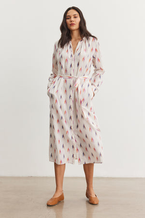 A person models the ALTA DRESS from Velvet by Graham & Spencer. The dress is white with a red and blue ikat weave pattern, featuring buttons and pockets. They complete the look with tan flats.