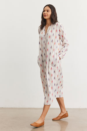 A person in the ALTA DRESS by Velvet by Graham & Spencer, featuring an ikat pattern of pointed shapes, strolls gracefully in brown flats on a plain floor.