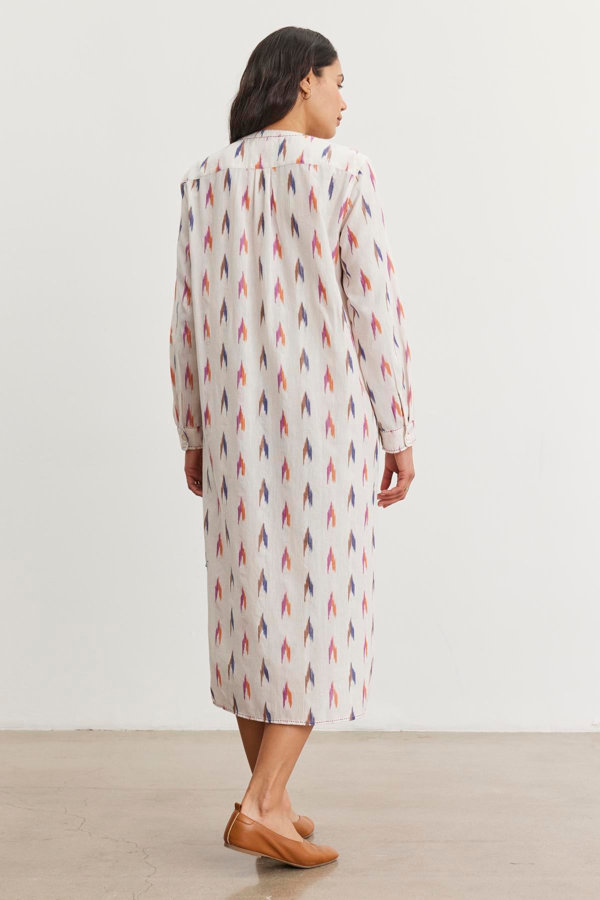 A person wearing Velvet by Graham & Spencer's ALTA DRESS, a long, patterned cotton dress with red and blue arrow designs and a detachable belt, stands back to the camera, paired with tan flats.