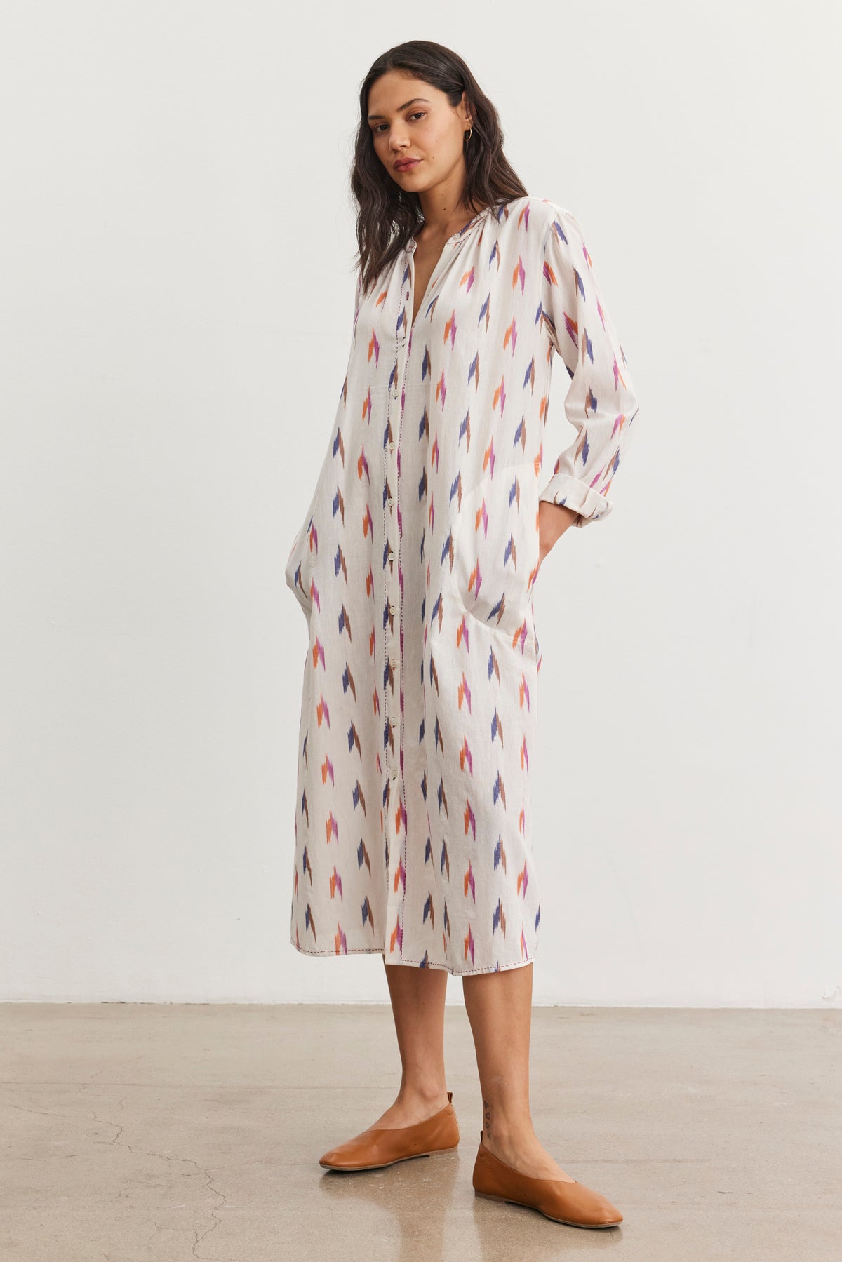 A person wearing the ALTA DRESS by Velvet by Graham & Spencer, a long patterned cotton dress with pockets and an intricate ikat weave, stands indoors on a plain floor.