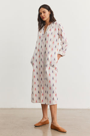 A person wearing the ALTA DRESS by Velvet by Graham & Spencer, a long patterned cotton dress with pockets and an intricate ikat weave, stands indoors on a plain floor.