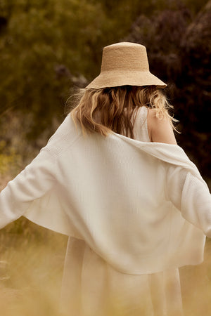 A woman with curly hair, seen from behind, wears a straw hat and a white flowing blouse in a sunlit, grassy field. She has draped over her shoulders a Velvet by Graham & Spencer Tava Cardigan.