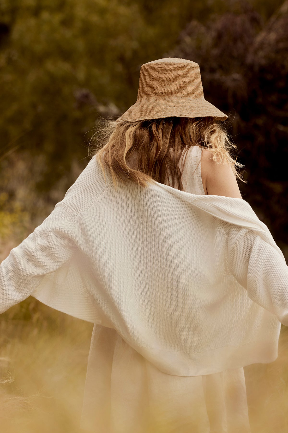 A person with long hair wears the CHIC CROCHET BUCKET HAT by Velvet by Graham & Spencer, paired with a white dress and cardigan, standing outdoors with their back to the camera and enjoying stylish sun protection.-39301115740353