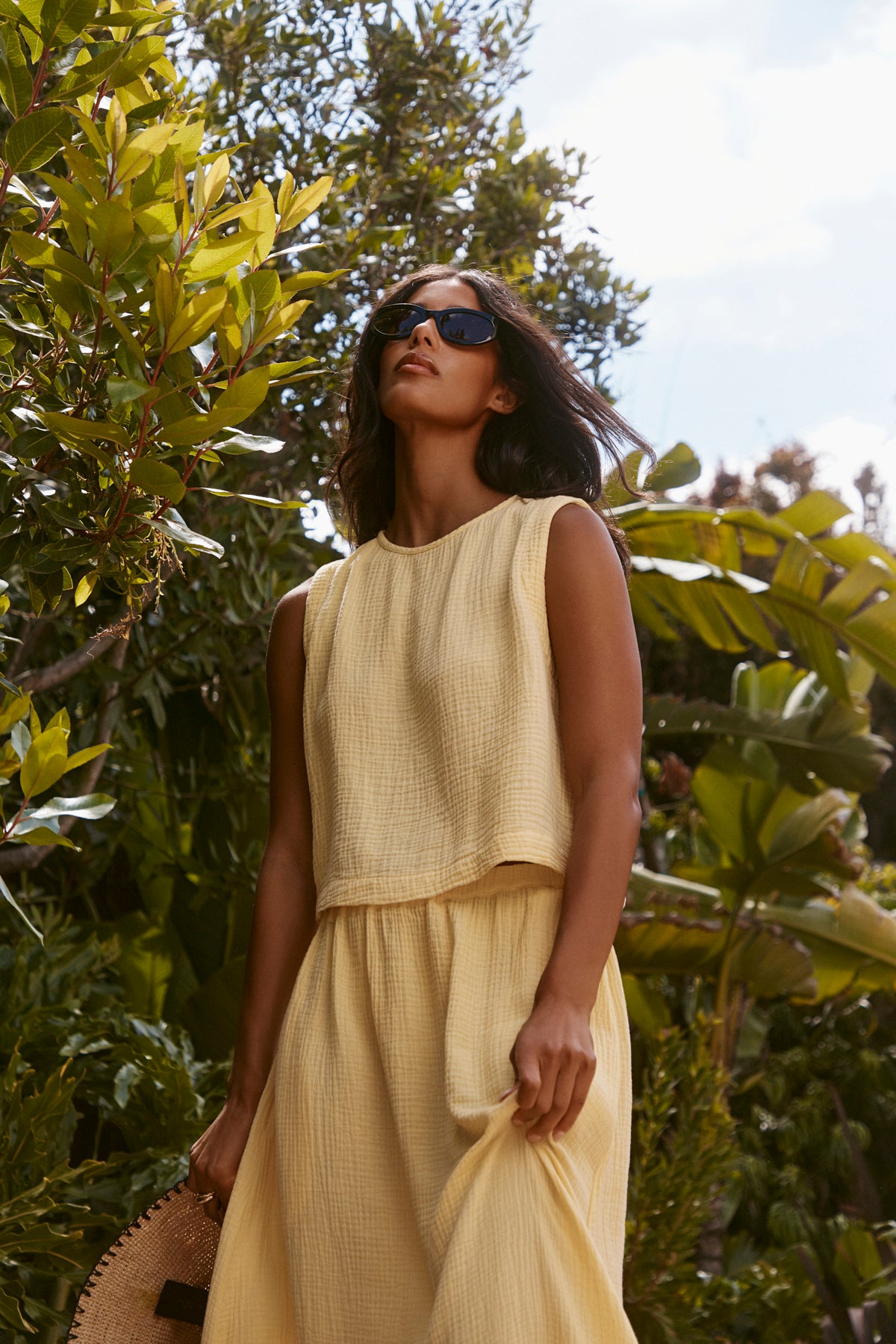A woman stands outdoors among green foliage, wearing sunglasses and the Velvet by Graham & Spencer CELESTE COTTON GAUZE SKIRT, styled with a sleeveless yellow top and straw hat—a breezy look for warm-weather days.