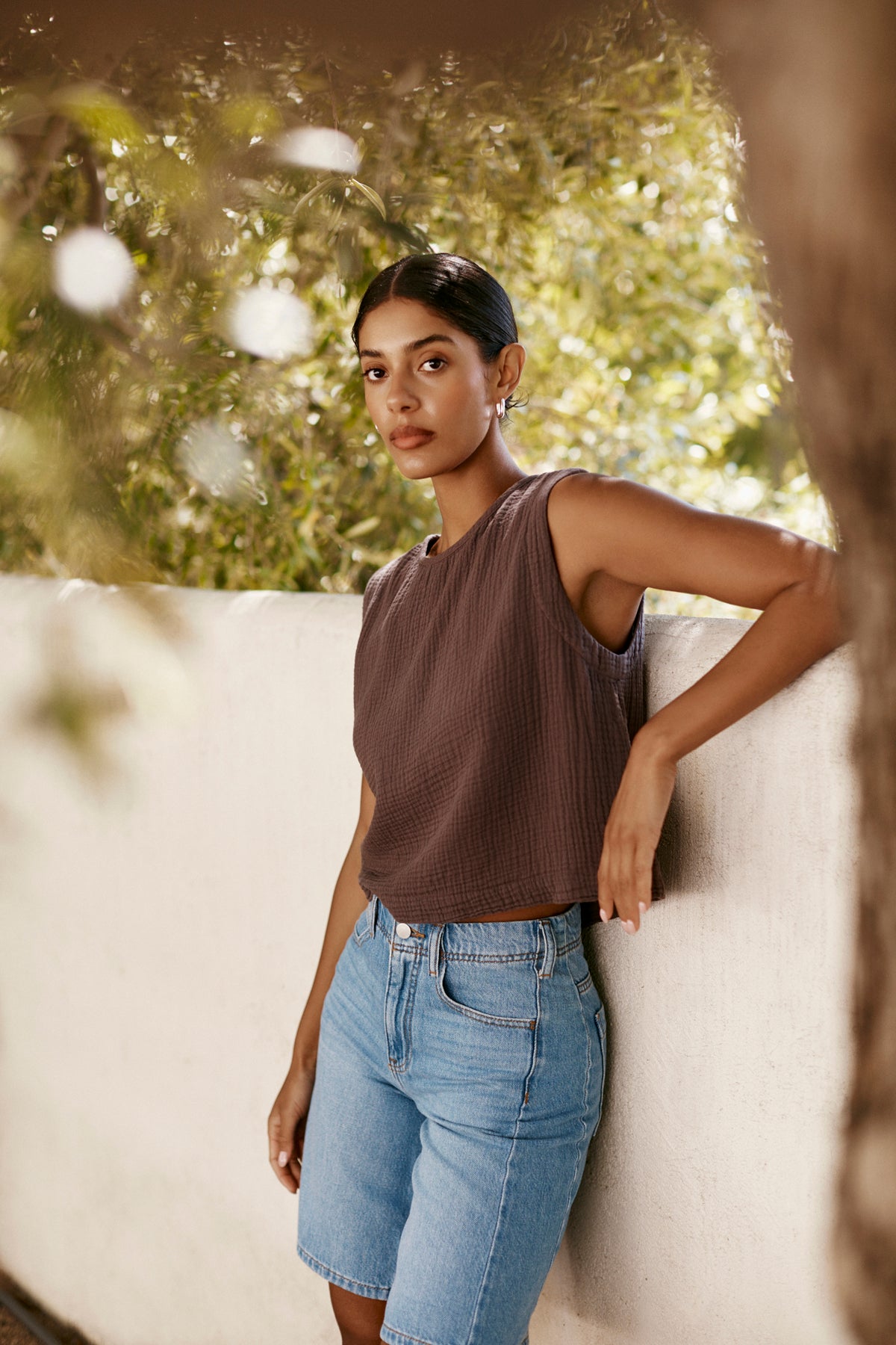 A woman with dark hair in a low bun poses outdoors by a white wall with greenery, wearing the MILNEY COTTON GAUZE TOP by Velvet by Graham & Spencer and denim shorts.-39266015379649
