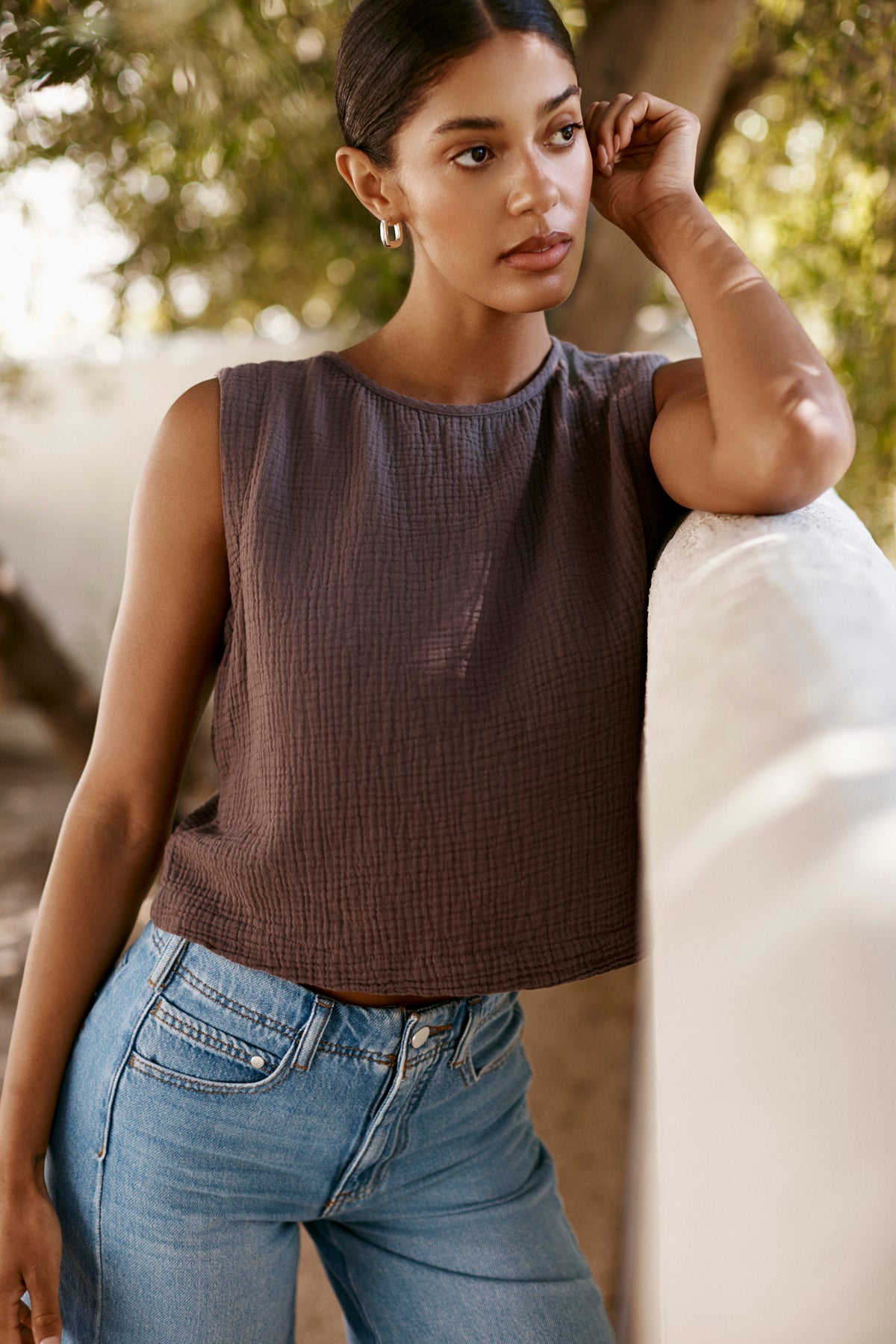 A woman stands outdoors, leaning on a white wall and wearing the MILNEY COTTON GAUZE TOP by Velvet by Graham & Spencer with blue jeans. She's framed by trees and natural light in the background.-39266015445185