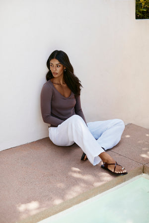 A woman relaxes on a patio by the pool, leaning against a white wall, wearing the Velvet by Graham & Spencer NOURA TEE in brown, paired with white pants and sandals.