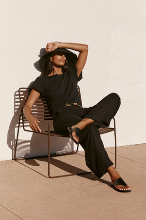 A woman in a black SOPHIA PANT by Velvet by Graham & Spencer, featuring a cropped balloon shape and elastic waist, sits outdoors in a wide-brim hat, shielding her eyes from the sun. This lightweight look is ideal for sunny days.