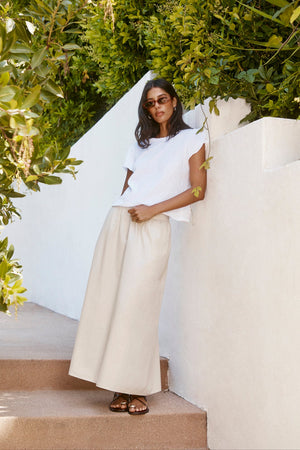 A woman in sunglasses, the PATRICIA TEE by Velvet by Graham & Spencer, and a beige maxi skirt stands on outdoor steps next to a white wall and green foliage.