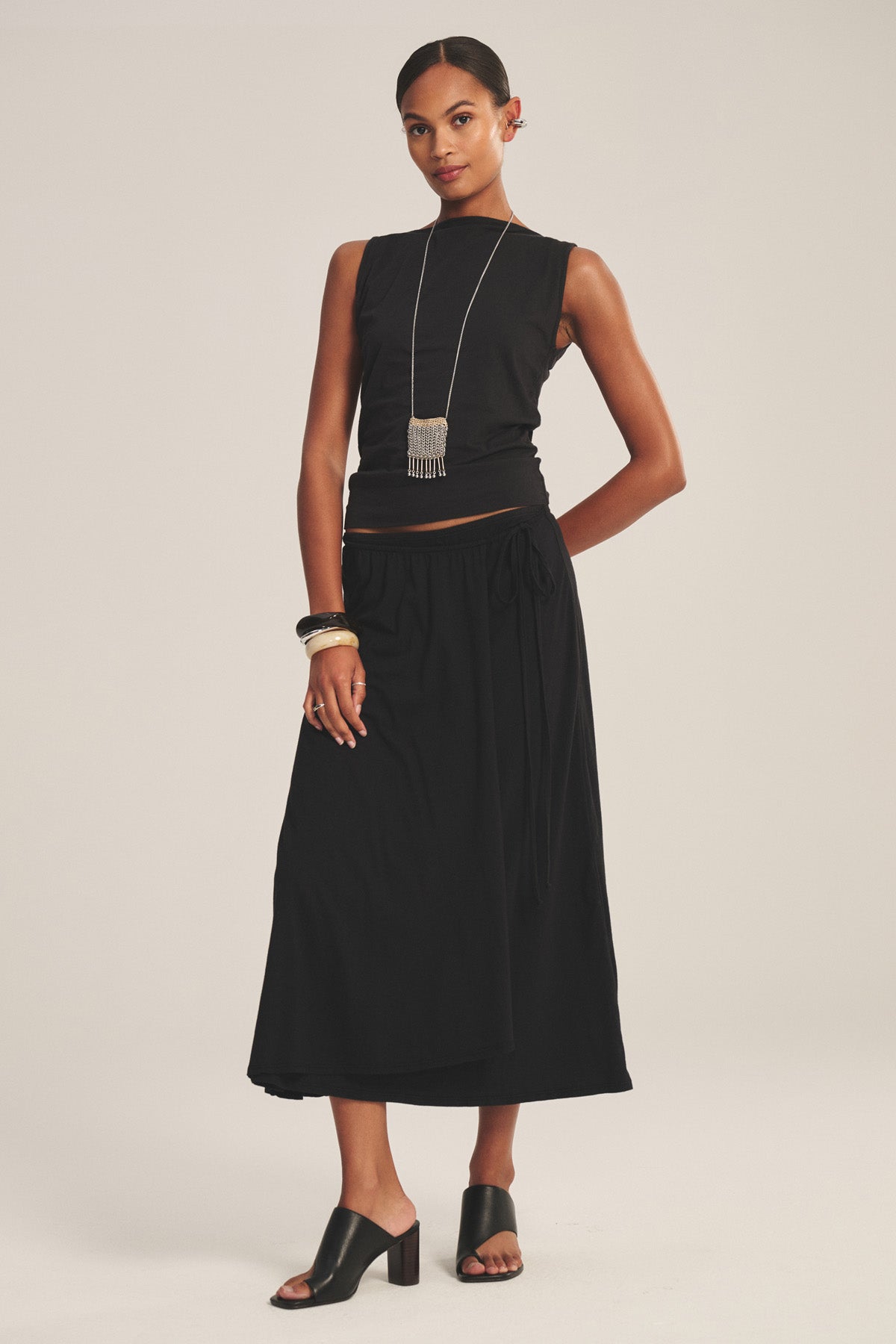 A woman stands against a plain light background wearing the IRENE SKIRT by Velvet by Graham & Spencer—a midi-length black cotton gauze wrap skirt—styled with a sleeveless black top, black heeled sandals, and silver jewelry.-41128085422273