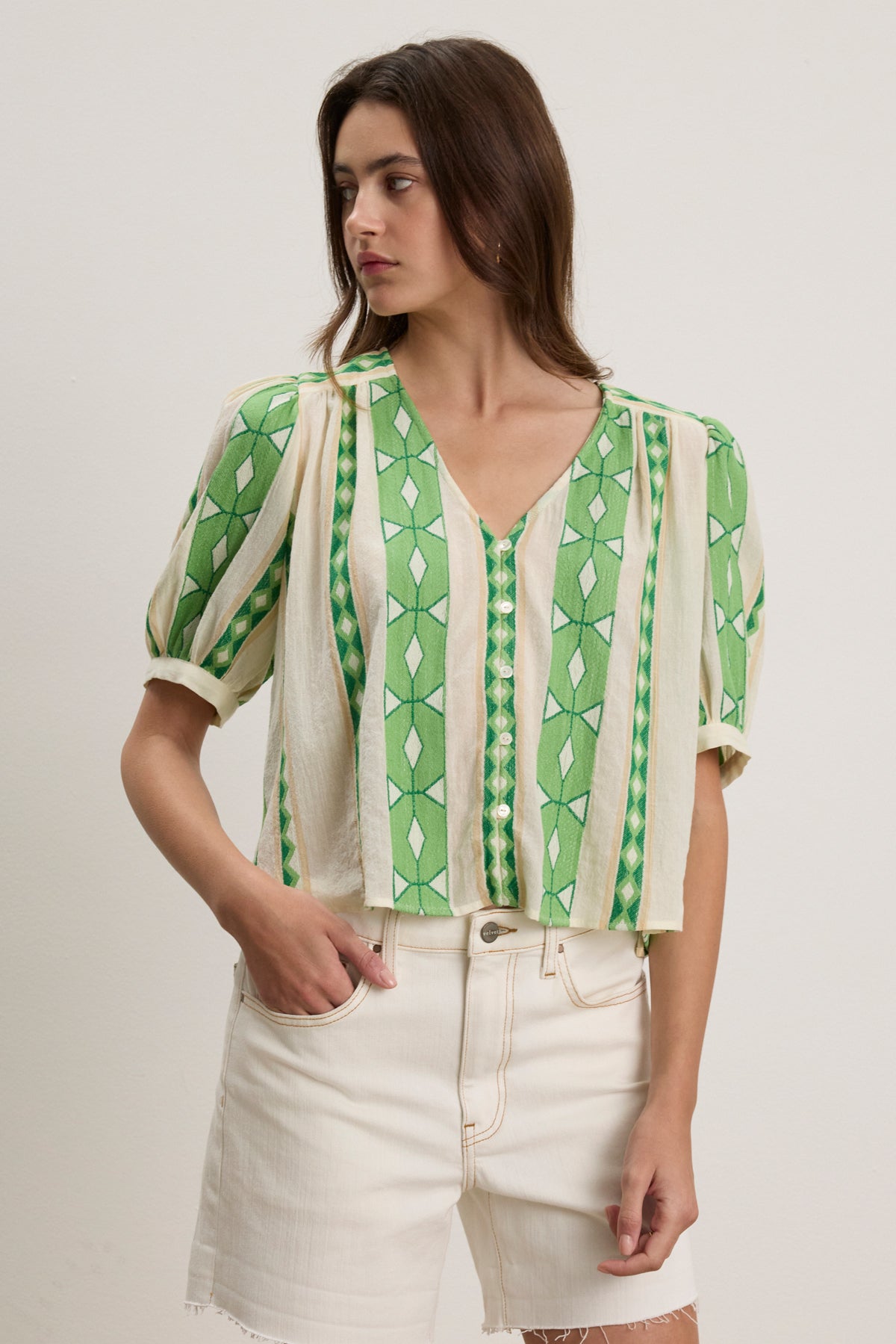 A woman models the MARISSA TOP by Velvet by Graham & Spencer—a green and cream patterned V-neck with short sleeves—paired with high-waisted white shorts, standing against a plain light background.