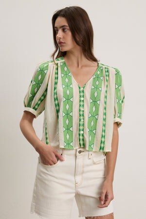 A woman models the MARISSA TOP by Velvet by Graham & Spencer—a green and cream patterned V-neck with short sleeves—paired with high-waisted white shorts, standing against a plain light background.