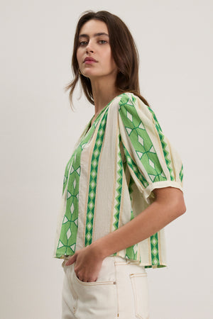 A person stands sideways against a plain background, wearing the MARISSA TOP by Velvet by Graham & Spencer—a green and cream cotton jacquard blouse—paired with high-waisted off-white jeans.