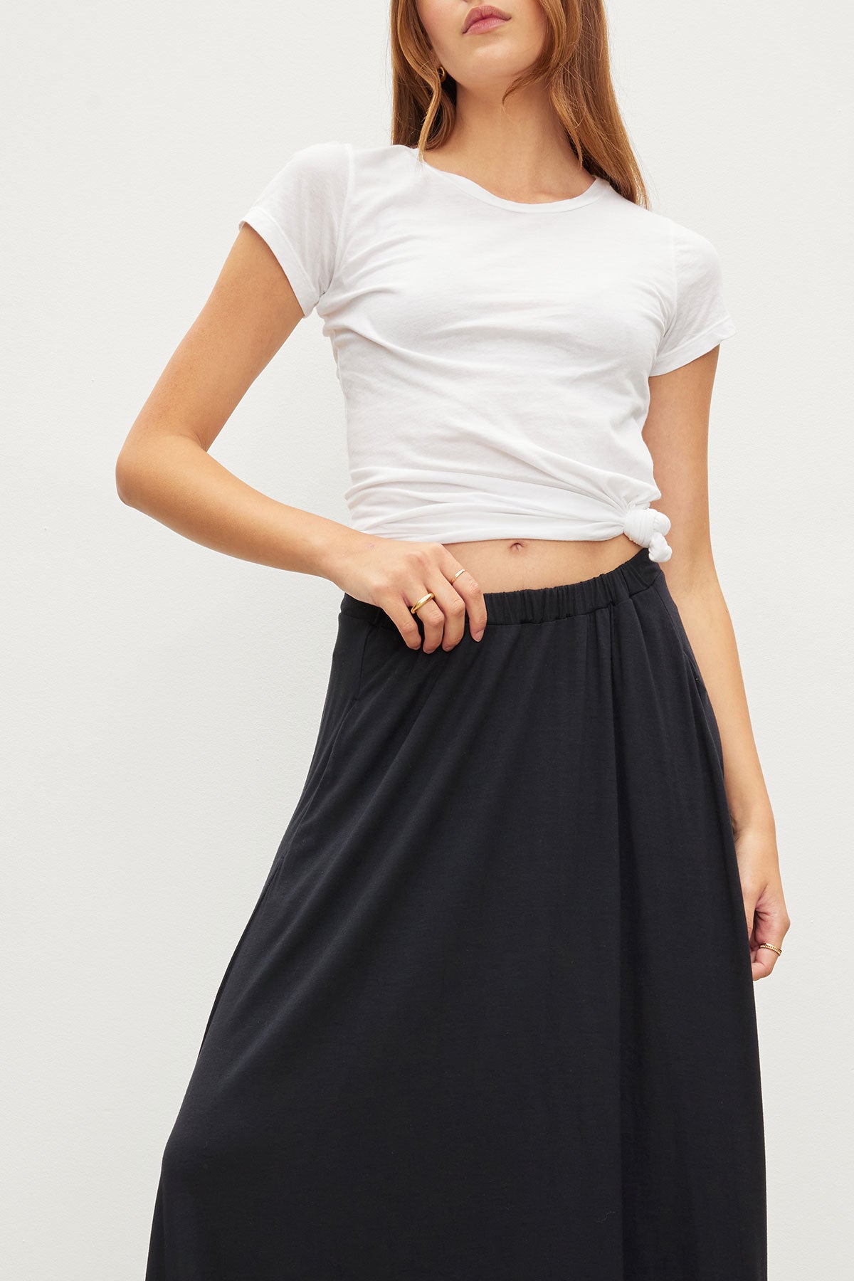 A woman stands against a plain light background wearing the JEMMA TEE by Velvet by Graham & Spencer, styled knotted at the waist with a high-waisted black skirt.-40101213503681