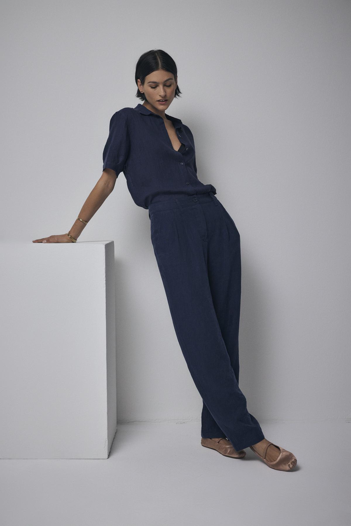 A woman in a CLAREMONT LINEN SHIRT from Velvet by Jenny Graham leans on a white cube in a minimalist setting.-36168678572225