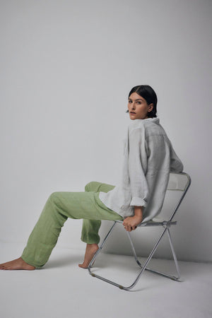 A woman sits on a metal chair against a plain background, wearing a white blouse and Velvet by Jenny Graham's PICO LINEN PANT in green, looking away thoughtfully.