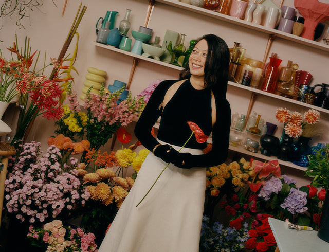 Woman standing in a flower shop with a variety of colorful flowers and ceramics on shelves.