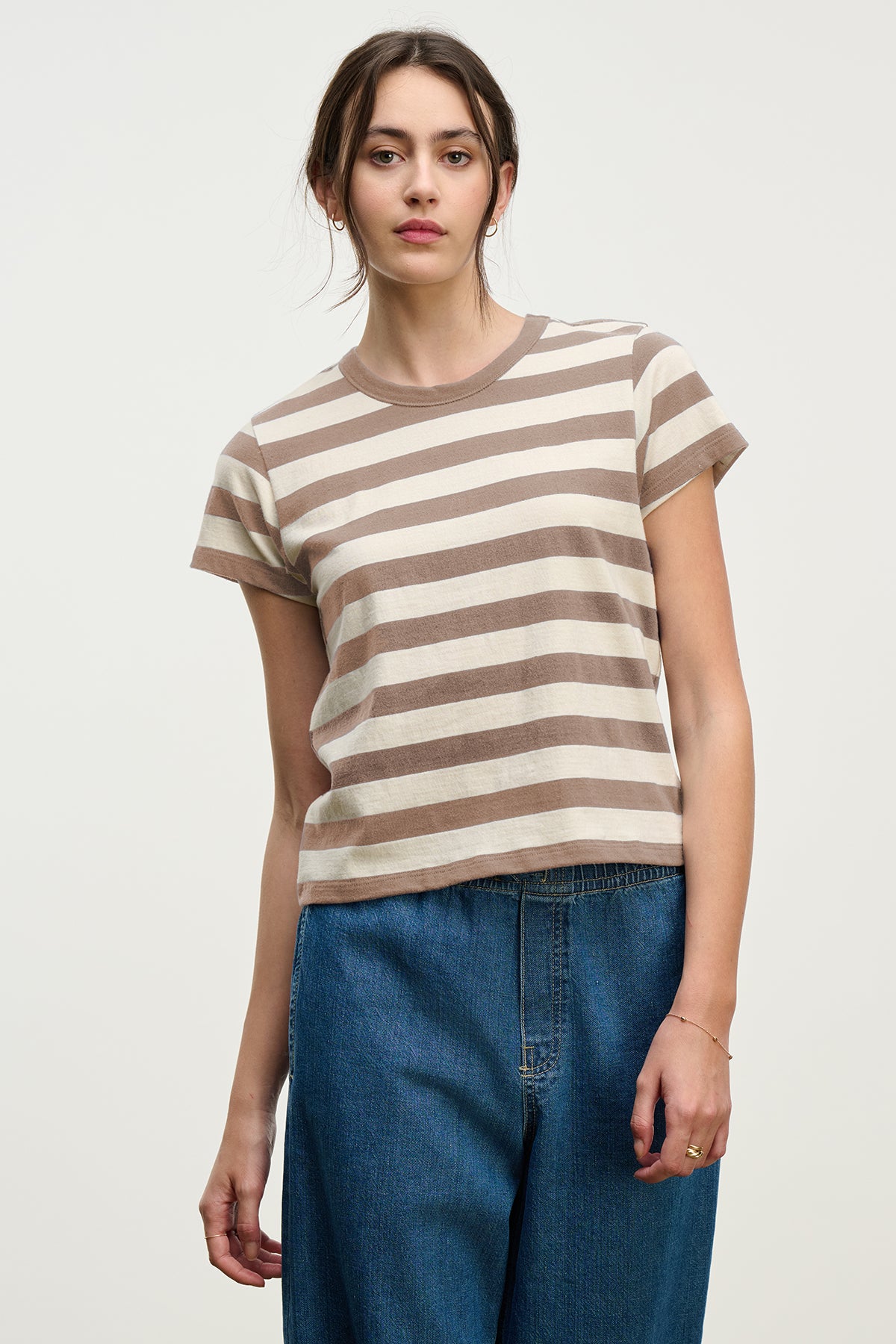 A woman faces forward in the ANAIS TEE by Velvet by Graham & Spencer—a short sleeve, crew neck cotton jersey with beige and brown stripes—paired with blue jeans against a plain light background.