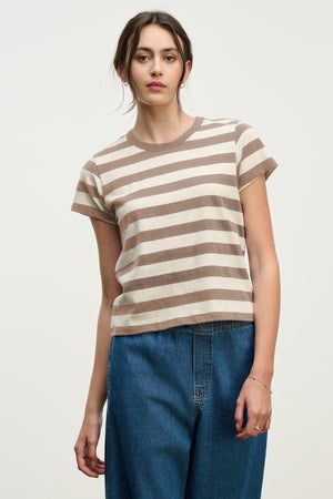A woman faces forward in the ANAIS TEE by Velvet by Graham & Spencer—a short sleeve, crew neck cotton jersey with beige and brown stripes—paired with blue jeans against a plain light background.