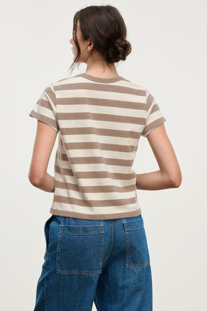 A woman with dark hair in a low bun is seen from the back, wearing the ANAIS TEE by Velvet by Graham & Spencer—a beige and white striped cotton jersey crewneck—paired with blue jeans against a plain light background.