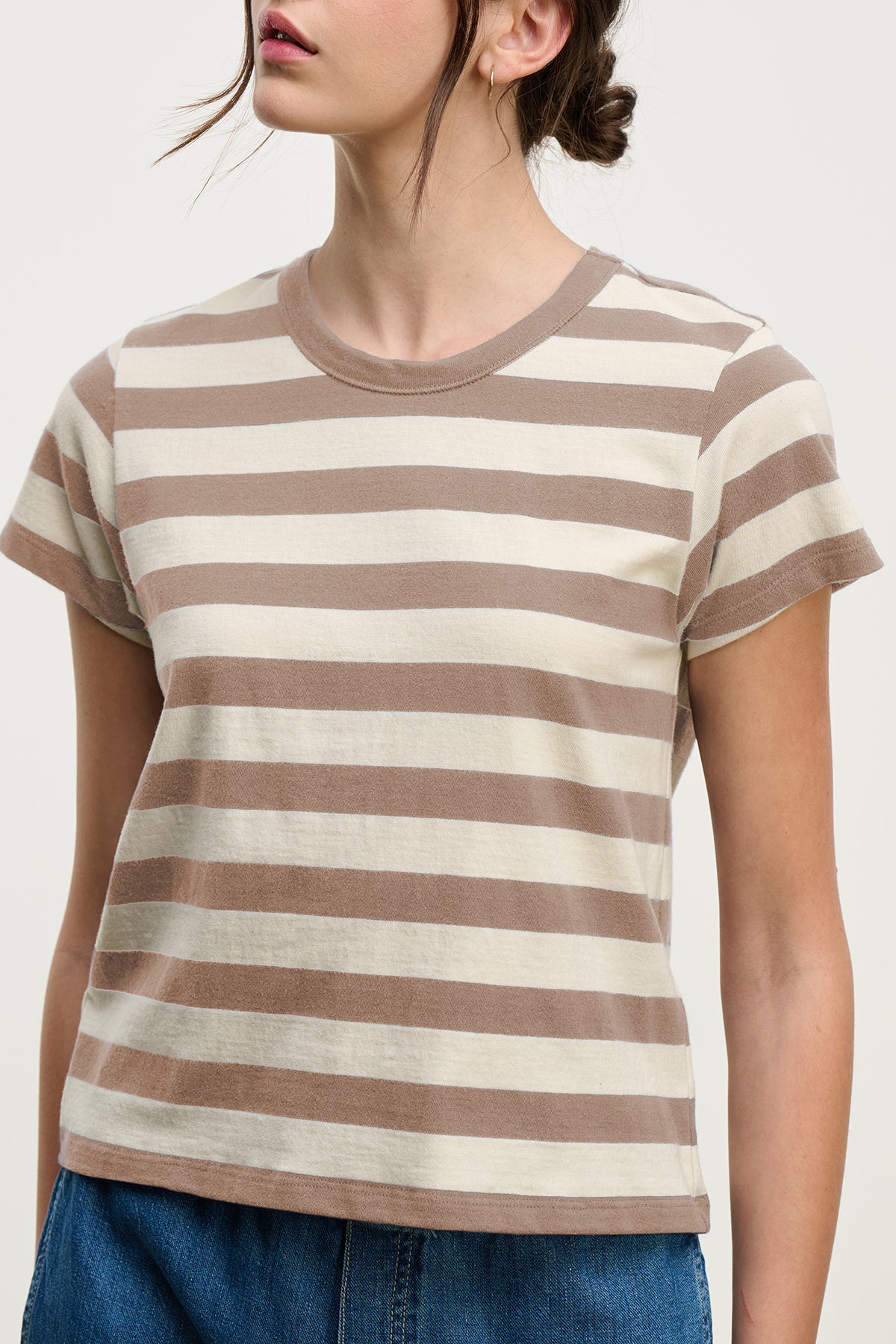 A woman in the ANAIS TEE by Velvet by Graham & Spencer, a short-sleeved beige and white striped cotton jersey crewneck, paired with blue jeans, stands against a plain background.