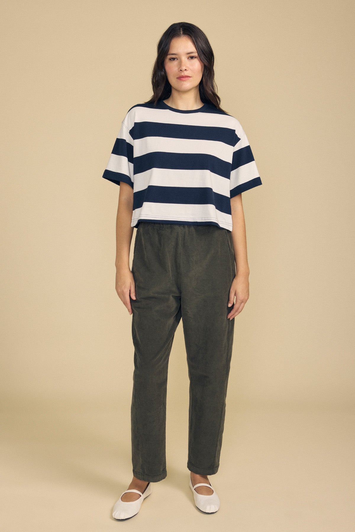A woman stands against a beige background wearing the FRYEE TEE by Velvet by Graham & Spencer—a striped cotton tee with a boxy fit—paired with olive green pants and white flat shoes.-41860431282369