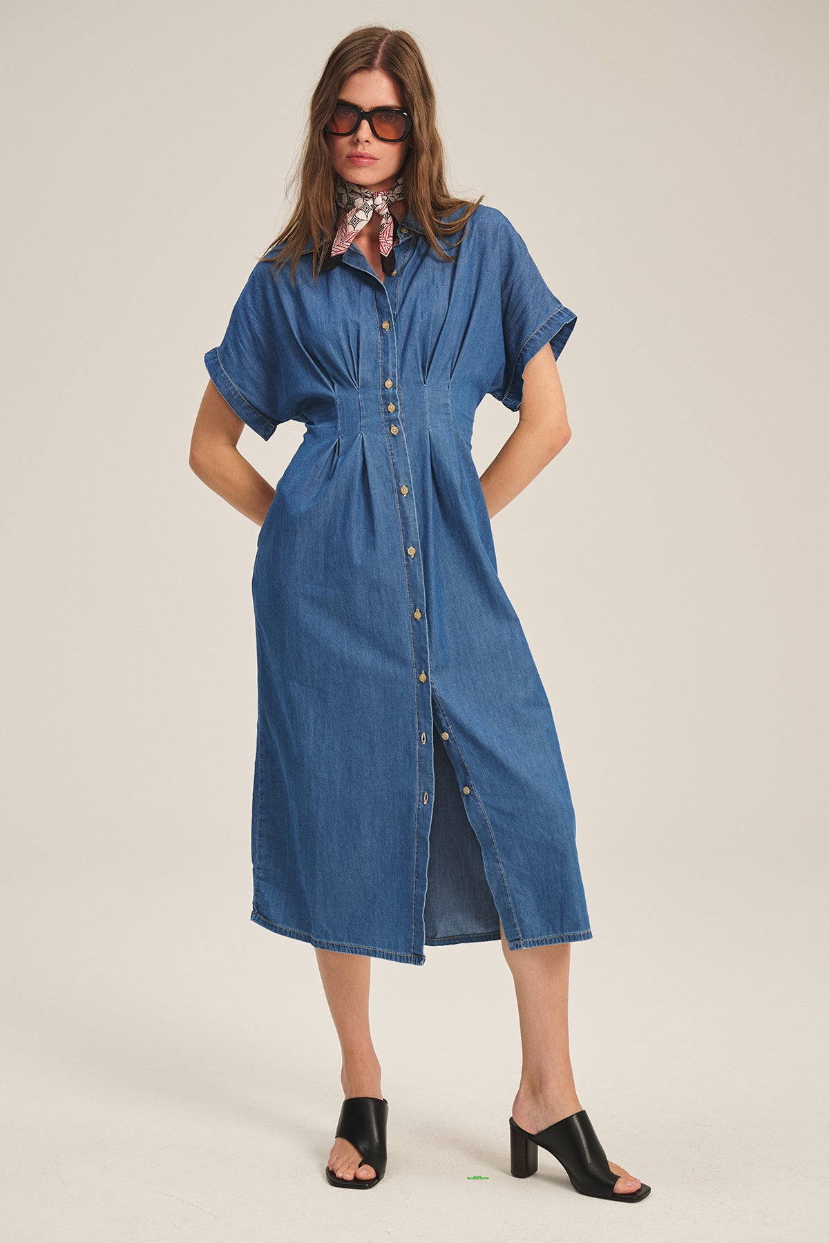 A model wears the Velvet by Graham & Spencer KARINA CHAMBRAY DRESS with short sleeves and pleats, styled with a pink scarf, black heeled mules, and large sunglasses against a plain background.-39600464527553