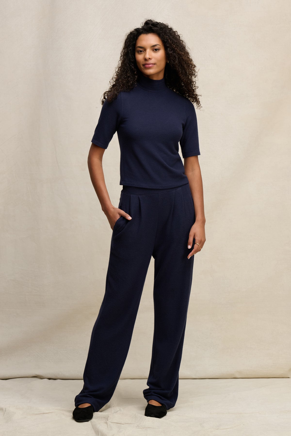 A woman stands against a neutral backdrop wearing Velvet by Graham & Spencer’s ELIYA SWEATPANT with a navy short-sleeve turtleneck top and black shoes.-39838349263041