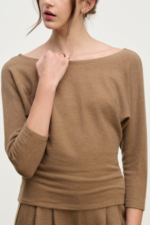 A woman wears the SELENA TOP by Velvet by Graham & Spencer—a light brown cotton viscose boatneck top with long sleeves and a relaxed fit—one hand on her collarbone, face partially out of frame.