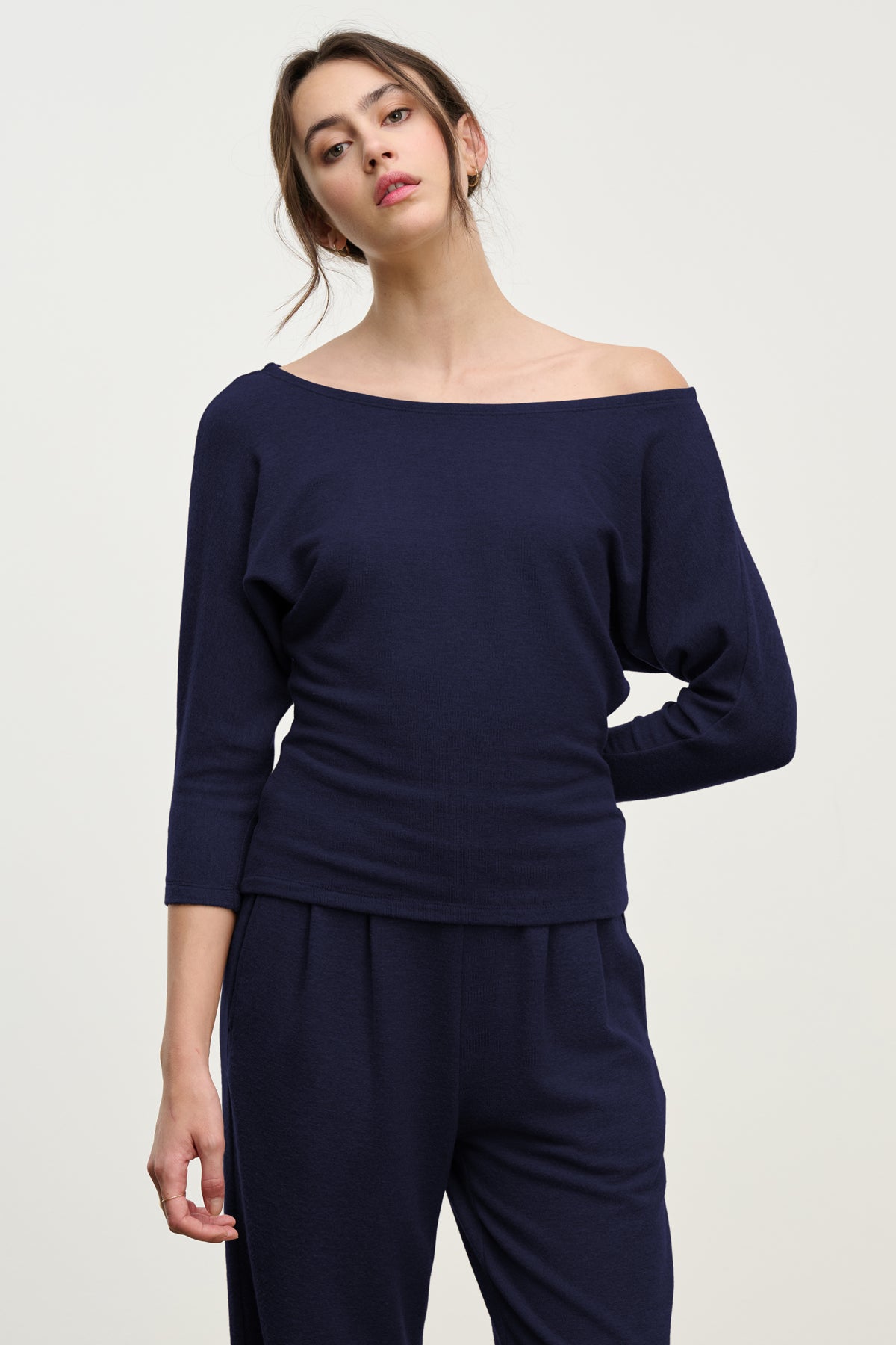 A woman stands against a plain background wearing the SELENA TOP by Velvet by Graham & Spencer in navy blue, featuring a boatneck and long sleeves, paired with matching pants.
