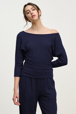 A woman stands against a plain background wearing the SELENA TOP by Velvet by Graham & Spencer in navy blue, featuring a boatneck and long sleeves, paired with matching pants.