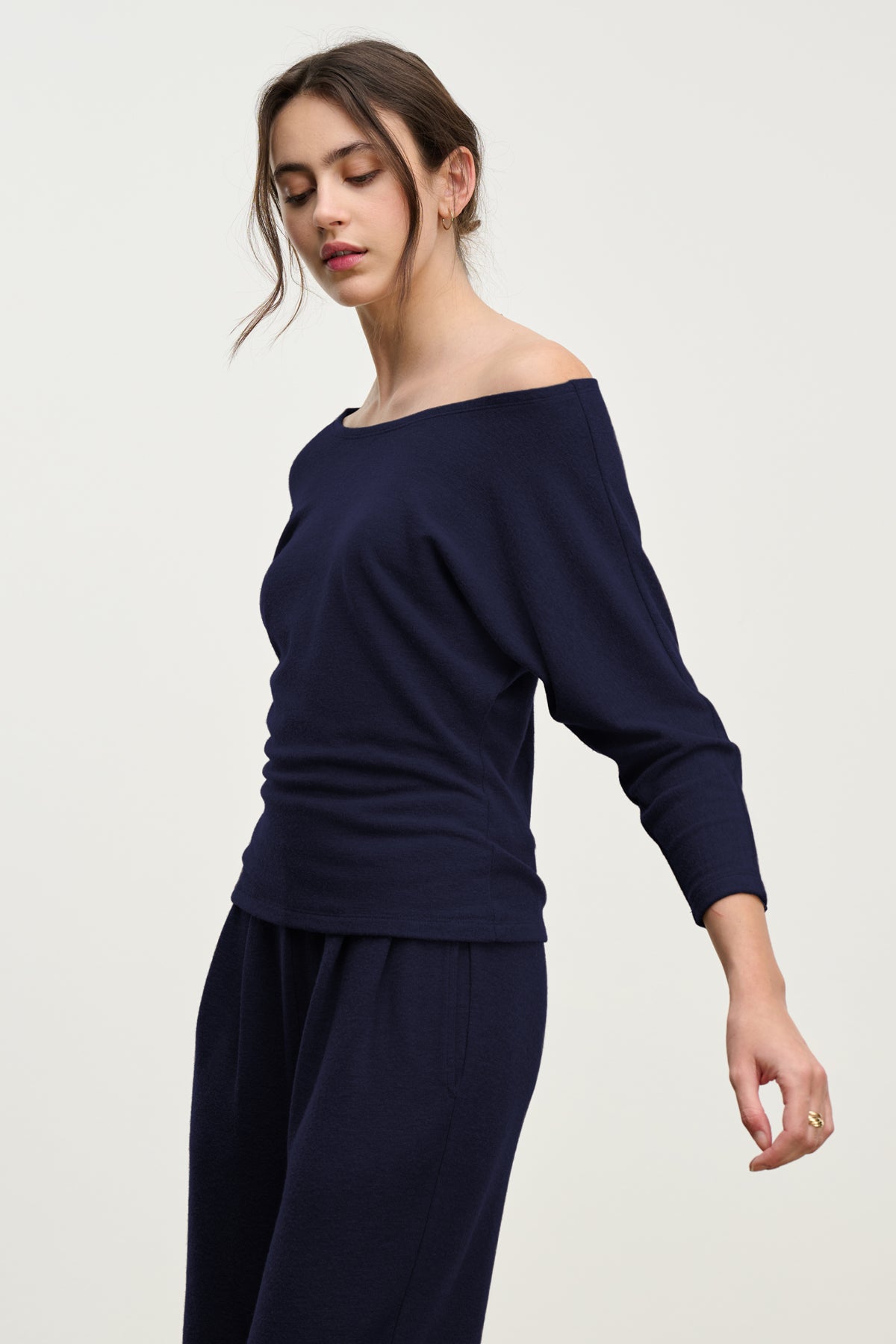 A woman in the SELENA TOP by Velvet by Graham & Spencer—navy blue, off-the-shoulder cotton viscose—paired with matching pants, stands against a light background, looking down with one arm slightly raised.-39814917914817