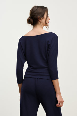 A woman stands with her back to the camera in a navy SELENA TOP by Velvet by Graham & Spencer, featuring a boatneck and dolman sleeves, paired with matching pants against a plain background.