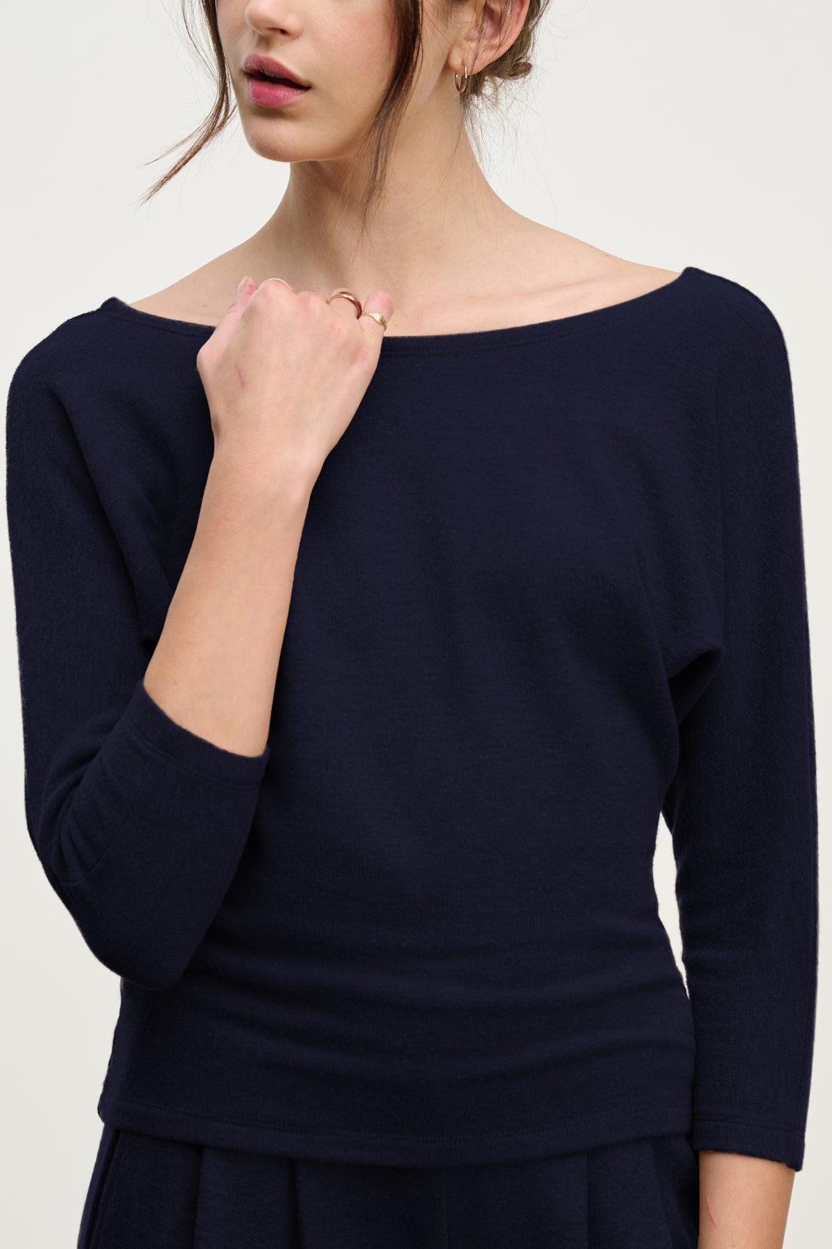 A woman with her hair tied back gently touches her collarbone while wearing the SELENA TOP by Velvet by Graham & Spencer—a navy blue, long-sleeved cotton viscose boatneck top.