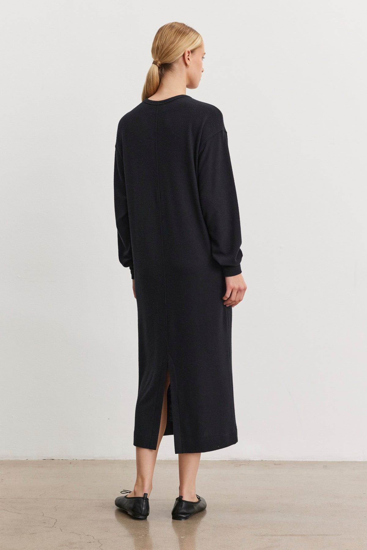 A person with blonde hair is wearing the AIDA DRESS by Velvet by Graham & Spencer, featuring a long black design with a crew neckline and side seam pockets. They are complemented by black shoes and stand on a concrete floor, facing away from the camera.-38436775755969