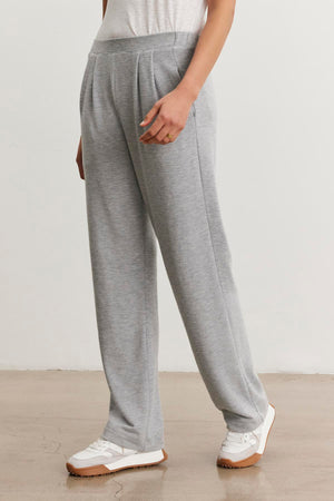 A person in Velvet by Graham & Spencer’s ELIYA SWEATPANT in ultra-soft light grey triblend knit, paired with a white t-shirt and sneakers, stands on a polished concrete floor against a plain white wall.