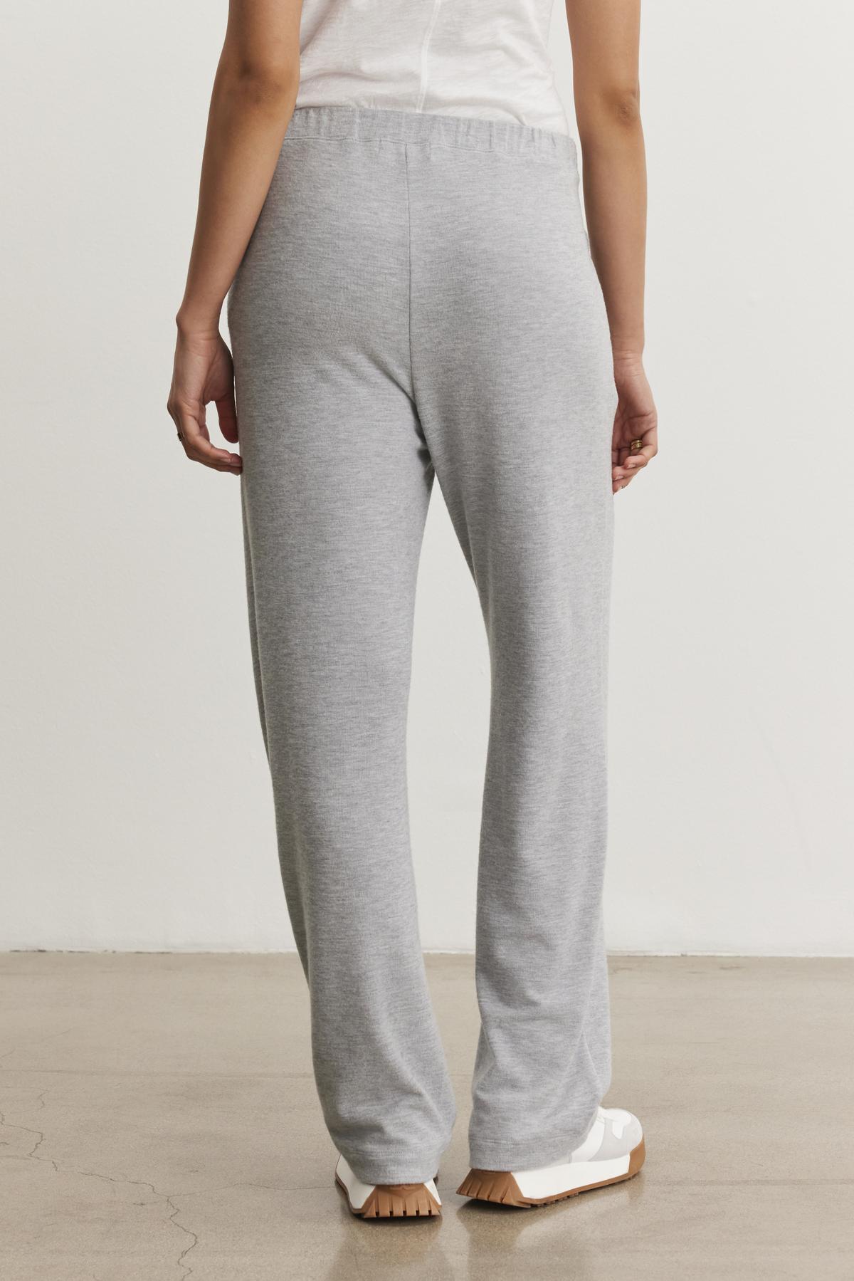 A person stands with their back to the camera, wearing Velvet by Graham & Spencer’s ELIYA SWEATPANT in ultra-soft light grey, paired with a white t-shirt and white shoes on a light-colored floor.-38433293533377