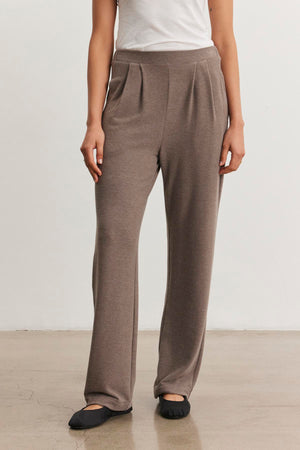 A person wears the ELIYA SWEATPANT by Velvet by Graham & Spencer in taupe with a white top and black flats, standing on a polished floor against a plain white wall.
