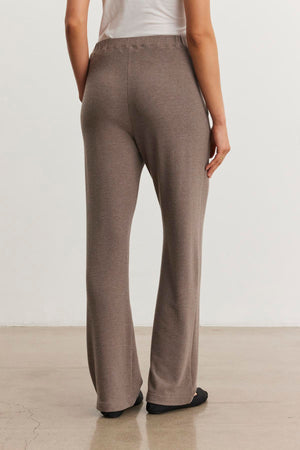 A person stands indoors facing away, wearing a white top and the ultra-soft ELIYA SWEATPANT by Velvet by Graham & Spencer in brown with black shoes on a smooth, light-colored floor.