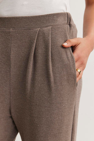 Close-up of a person wearing Velvet by Graham & Spencer’s ELIYA SWEATPANT in ultra-soft brown triblend knit with pleats, paired with a white top and gold ring, one hand tucked in the pocket.