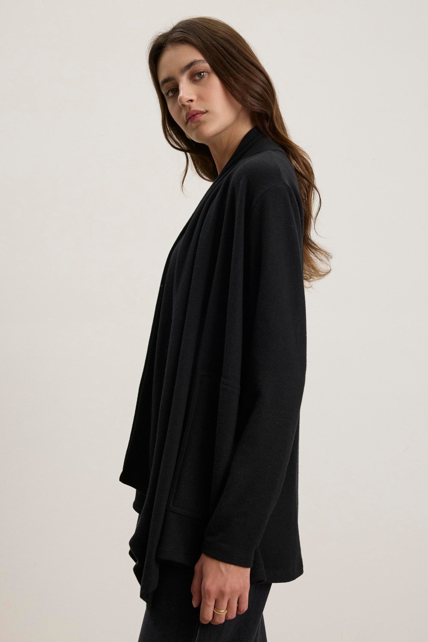 A person with long hair is wearing the LISA CARDIGAN by Velvet by Graham & Spencer, a black triblend knit cardigan with an open front and side patch pockets, standing against a plain background.-38433284423873
