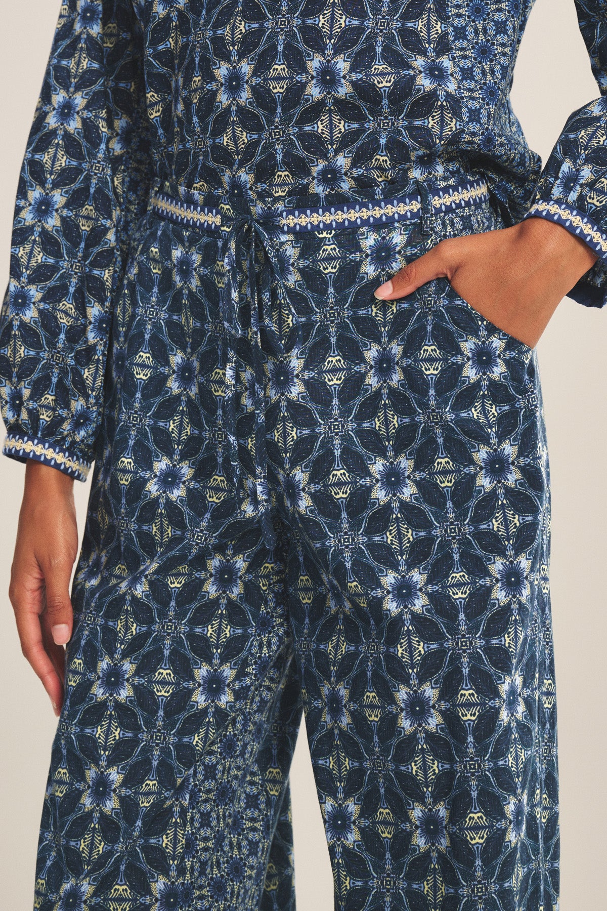 A person wears the Velvet by Graham & Spencer KENDRA PANT, a blue patterned lightweight cotton pant with a drawstring waist, geometric and floral designs, and a straight-leg silhouette.-41008281911489
