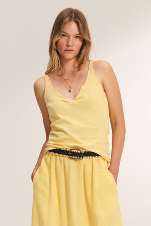 Woman in the KIM TANK TOP by Velvet by Graham & Spencer—a yellow lightweight cotton tank with a scoop neckline and matching skirt, accessorized with layered necklaces and a black belt, stands against a neutral background.