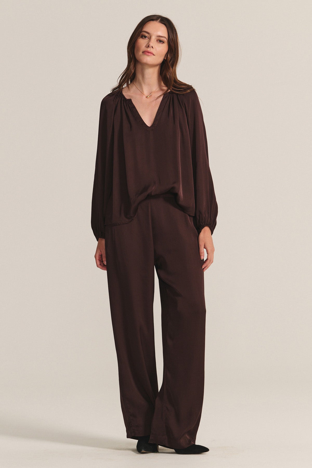 A woman stands against a plain background, wearing a loose-fitting, long-sleeved dark brown blouse and the LARKIN PANT by Velvet by Graham & Spencer—wide-leg satin pants paired with black shoes for effortless everyday style.-40248086921409