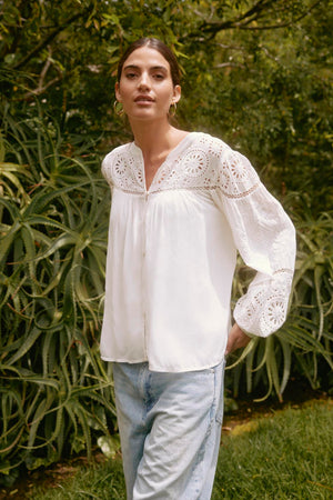 A person wearing the LIBBY TOP from Velvet by Graham & Spencer and jeans stands outdoors in front of greenery.