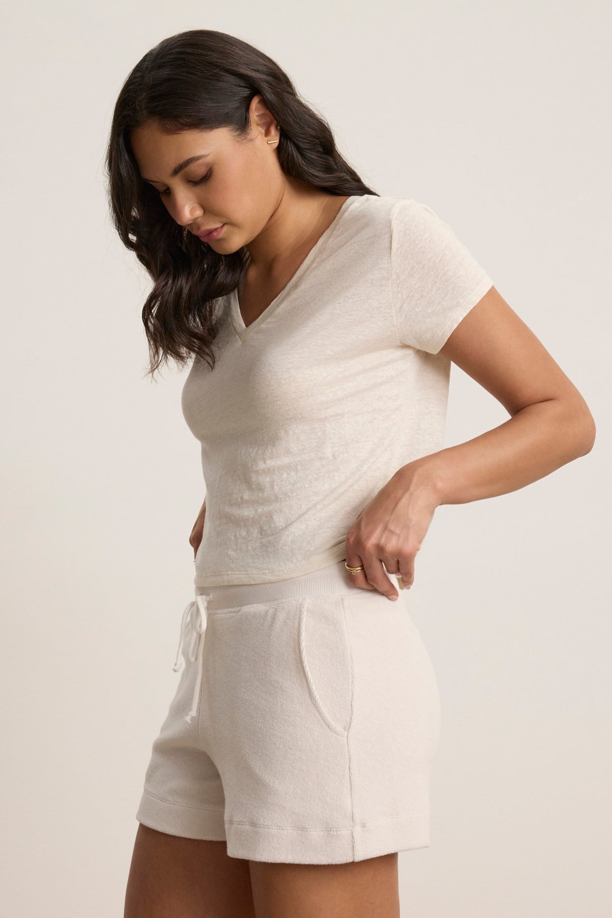 A woman in the AMBER LINEN KNIT TEE by Velvet by Graham & Spencer, a light-colored, cropped V-neck shirt paired with matching drawstring shorts, stands looking down and adjusts her shorts, capturing effortless casual comfort.-39123370344641