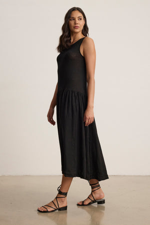 A woman stands indoors wearing the MABLE LINEN KNIT DRESS by Velvet by Graham & Spencer with black strappy sandals, her hair down as she looks slightly to the side.