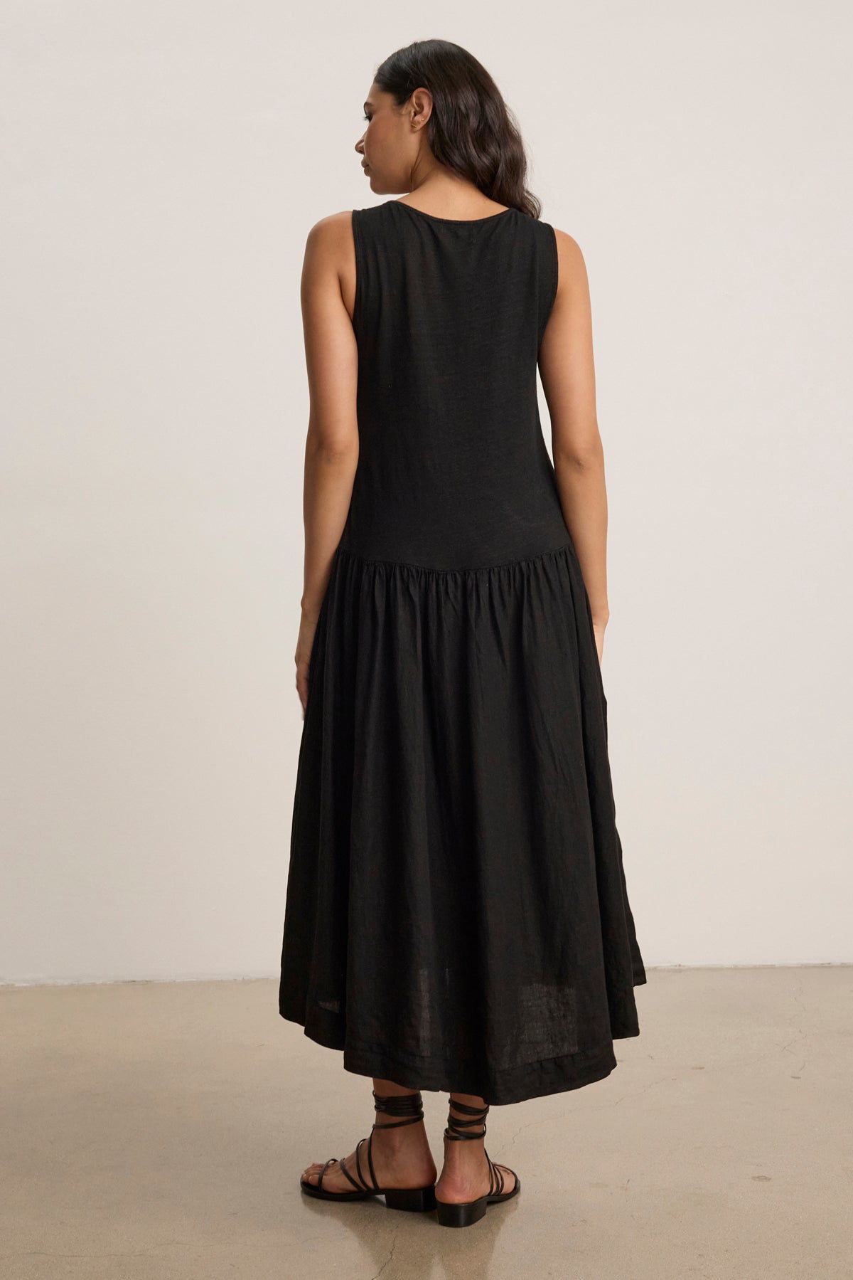 A woman stands indoors, facing away from the camera, in the MABLE LINEN KNIT DRESS by Velvet by Graham & Spencer—a sleeveless black linen blend midi dress—paired with black sandals against a plain background.