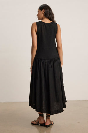 A woman stands indoors, facing away from the camera, in the MABLE LINEN KNIT DRESS by Velvet by Graham & Spencer—a sleeveless black linen blend midi dress—paired with black sandals against a plain background.