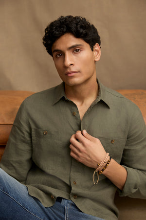 A young man with short curly hair wears the LINDEN LINEN BUTTON-UP SHIRT by Velvet by Graham & Spencer in olive green and blue jeans, sitting on a brown couch and looking at the camera with a neutral expression.